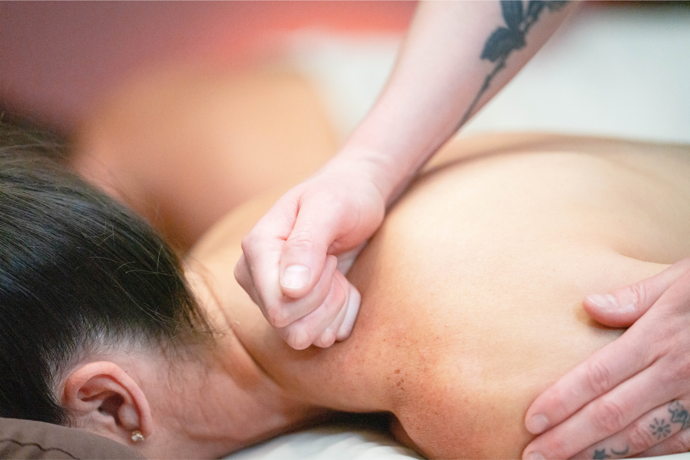 A person's bare back and shoulder receiving a massage. One hand uses a fist to knead the shoulder, while another hand supports. The person lies face down.