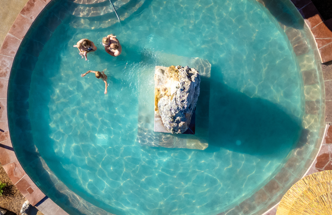 Three people swim in a circular pool with turquoise water. A natural rock fountain sprays water, surrounded by flagstone paving and two thatched umbrellas.