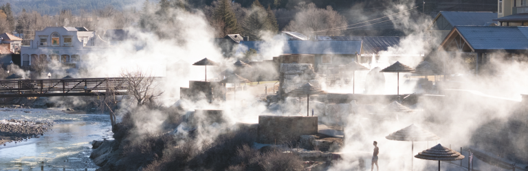 Steaming tiered hot spring pools with people relaxing in misty waters, alongside a river, bridge, and buildings, backed by pine forests and snowy mountains.