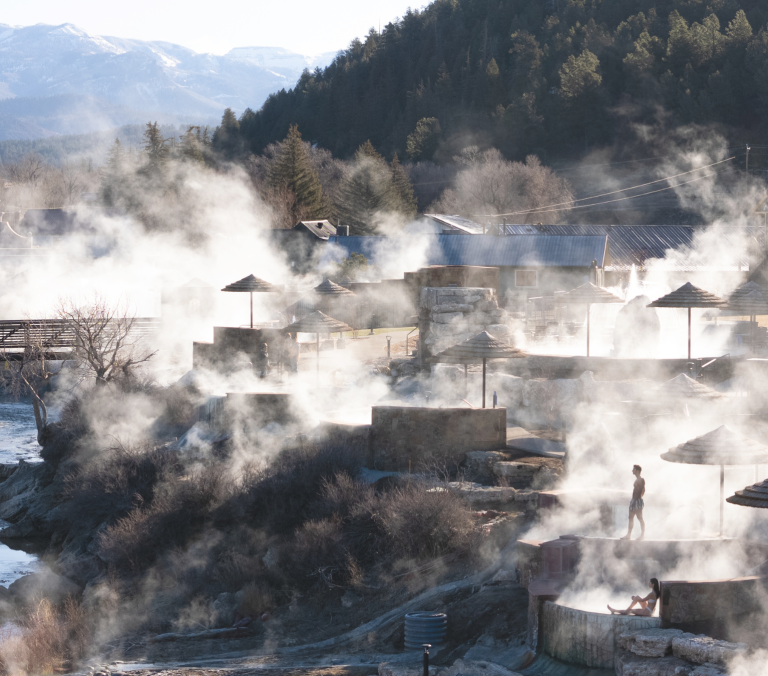 Steaming tiered hot spring pools with people relaxing in misty waters, alongside a river, bridge, and buildings, backed by pine forests and snowy mountains.