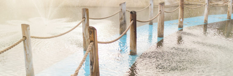Wooden posts linked by ropes form a fence extending across rippling water. A light blue submerged pathway lies beneath, with a misty fountain left and snow-dusted rocks right.