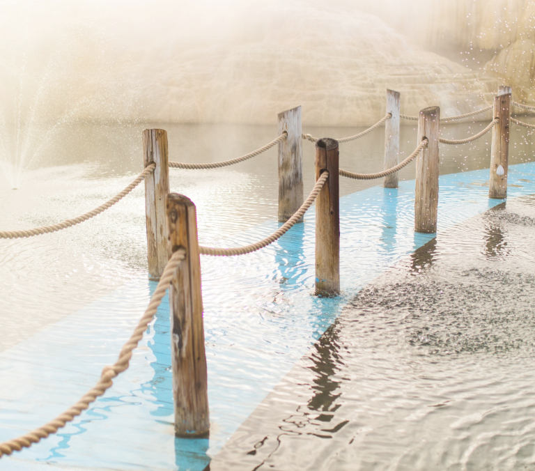 Wooden posts linked by ropes form a fence extending across rippling water. A light blue submerged pathway lies beneath, with a misty fountain left and snow-dusted rocks right.