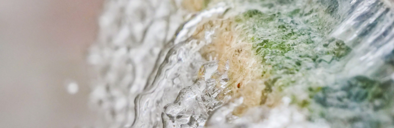 Water cascades downwards over a textured surface with green, beige, and white tones, creating ripples and droplets.