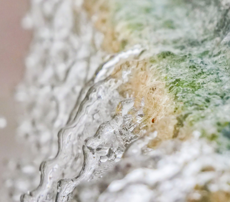 Water cascades downwards over a textured surface with green, beige, and white tones, creating ripples and droplets.