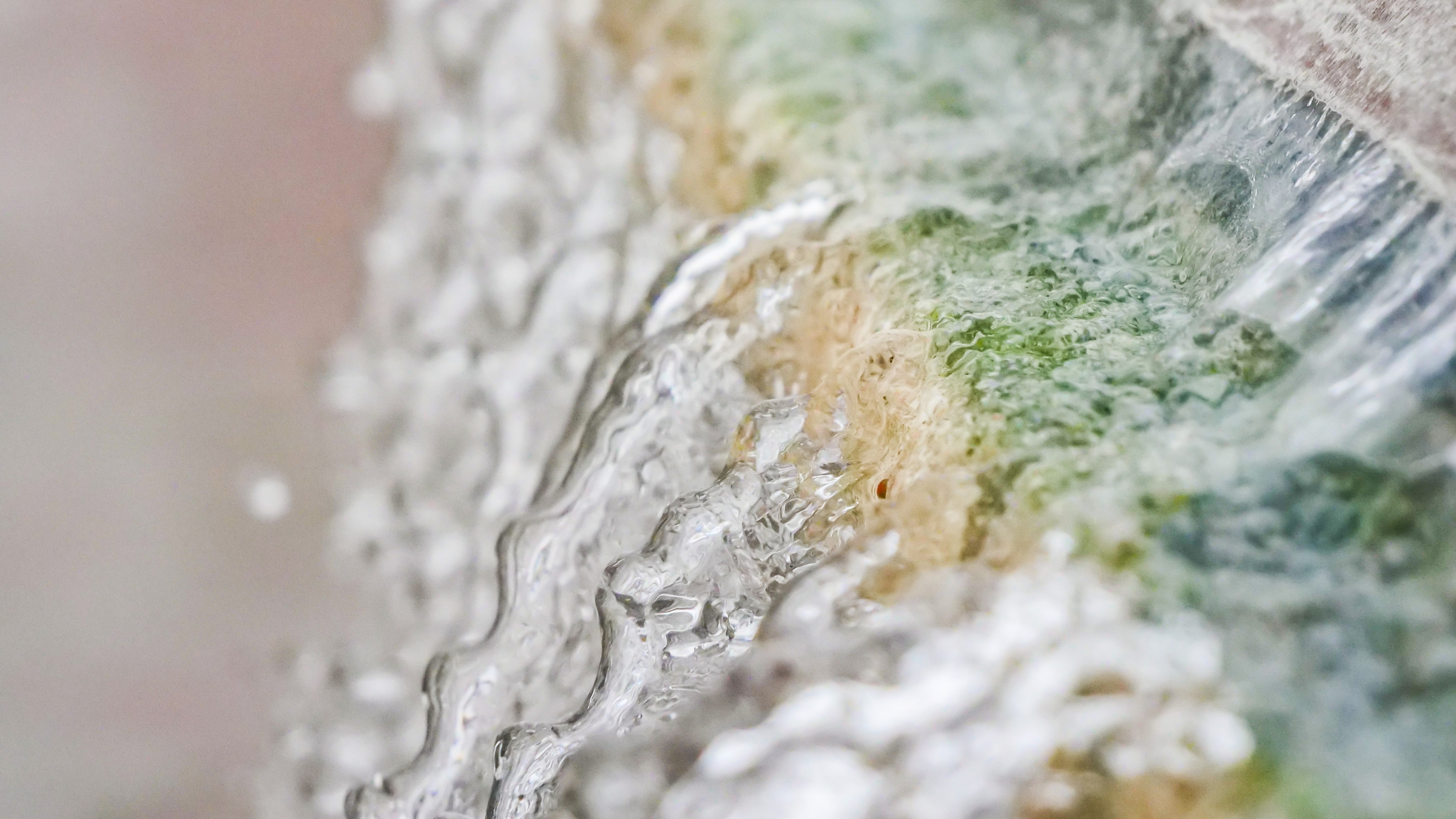 Water cascades downwards over a textured surface with green, beige, and white tones, creating ripples and droplets.