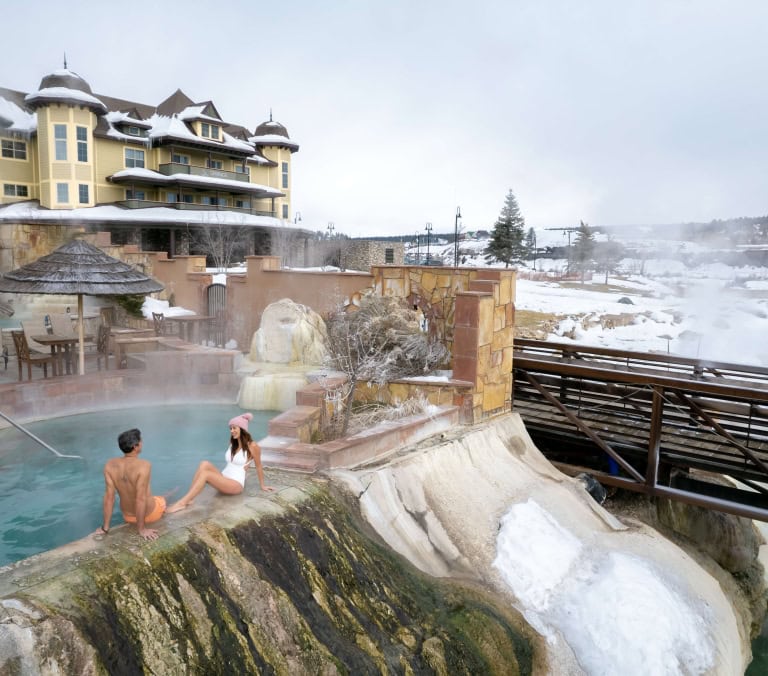 A couple relaxes in a steaming outdoor hot spring pool, next to a large yellow hotel with snow on the roof. The snowy landscape features distant buildings and trees. Text: SPRINGS