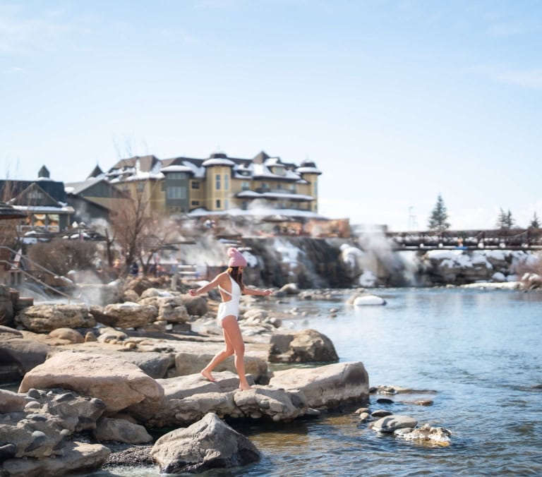 A woman in a white swimsuit and pink hat walks barefoot on rocks beside a steaming river. Snow-covered banks and a large hotel with a 