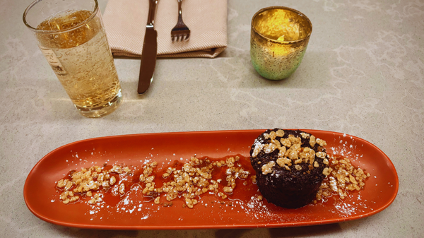 A chocolate lava cake, garnished with nuts, powdered sugar, and syrup, sits on an orange plate. A bubbly drink, silverware, and lit candle are beside it on a light gray counter.
