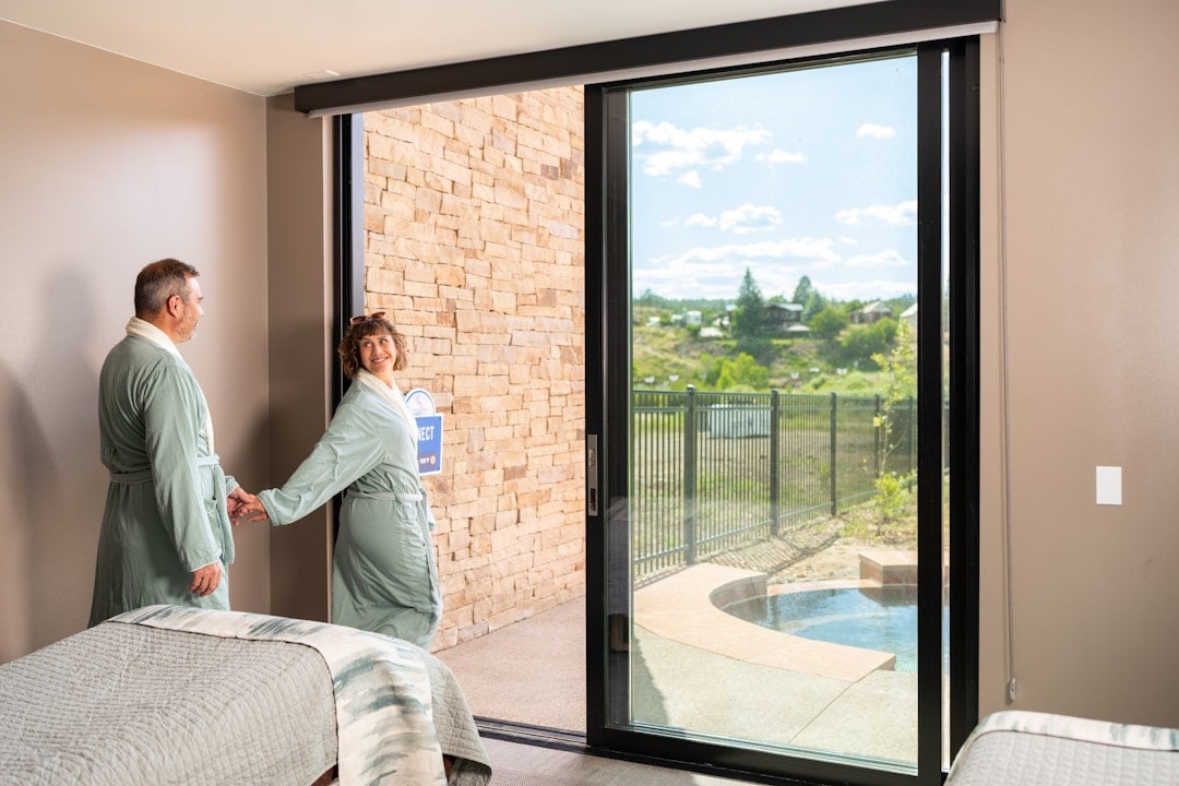 A smiling couple in bathrobes holds hands by a sliding door opening to an outdoor hot tub patio and distant houses on a hill. Text: CONNECT.