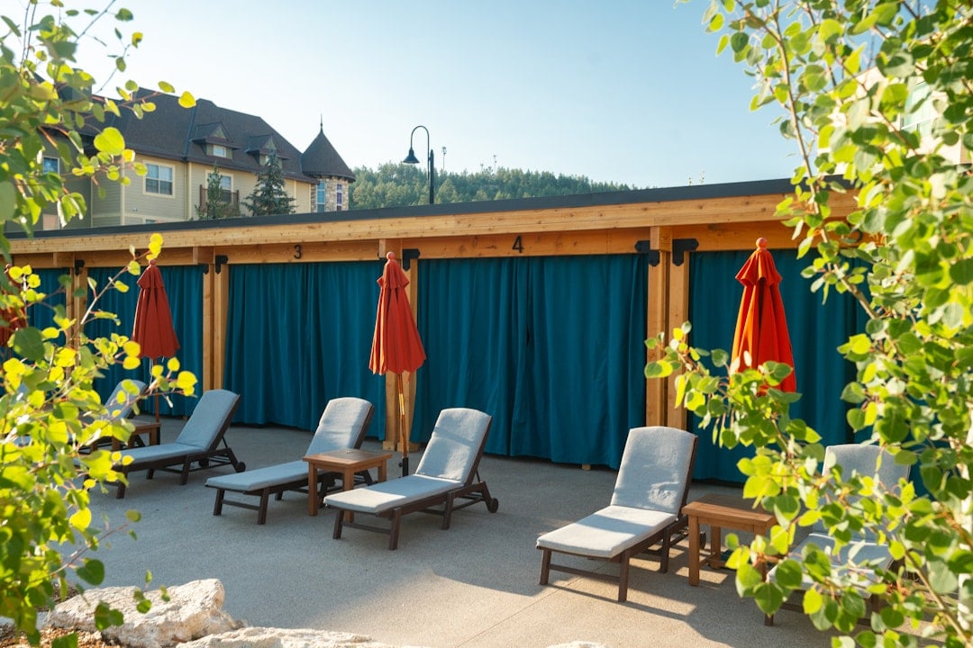 Lounge chairs and closed orange umbrellas sit on a sunny patio with cabanas featuring teal curtains marked **3** and **4**. A resort building is in the background.