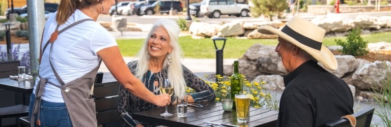 A server delivers wine to a smiling elderly woman at an outdoor patio table where an elderly man also sits, with buildings and mountains in the background.