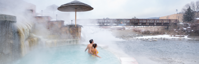 A couple embraces in a steamy hot spring pool by a waterfall, with a snowy river and a bridge bearing 