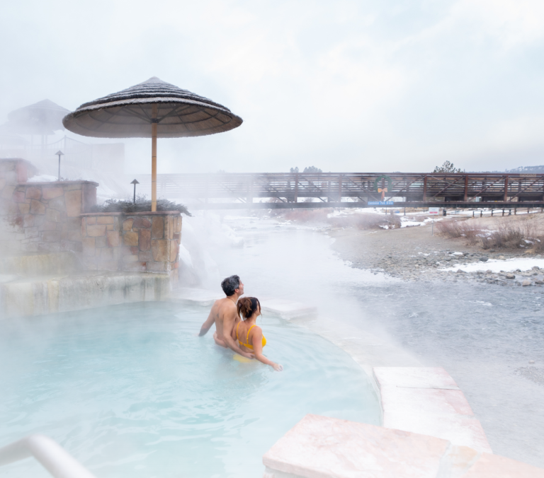 A couple embraces in a steamy hot spring pool by a waterfall, with a snowy river and a bridge bearing 