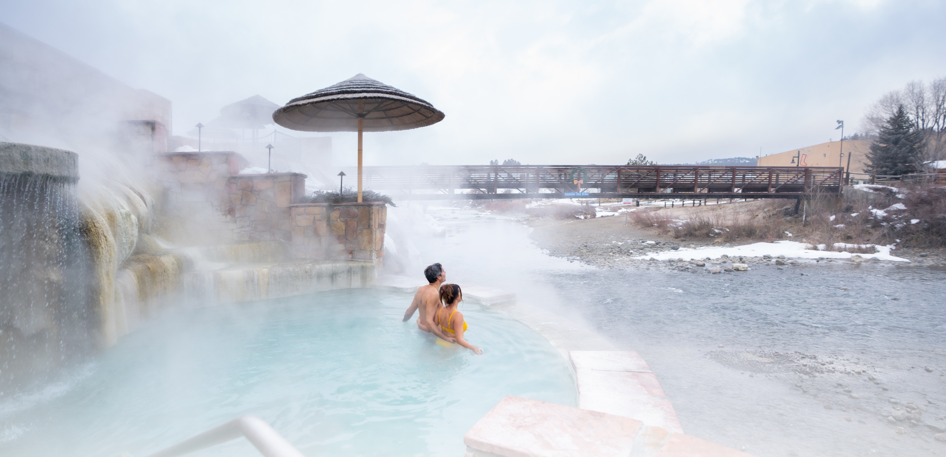 A couple embraces in a steamy hot spring pool by a waterfall, with a snowy river and a bridge bearing 