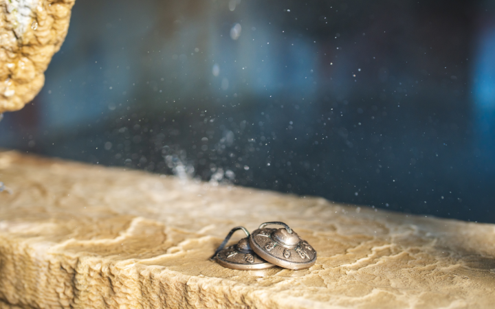 Two ornate, bronze-colored circular objects, linked by a black cord, rest on a light brown, rough stone surface. Water drips from above, creating a fine mist against a blurred dark blue background.