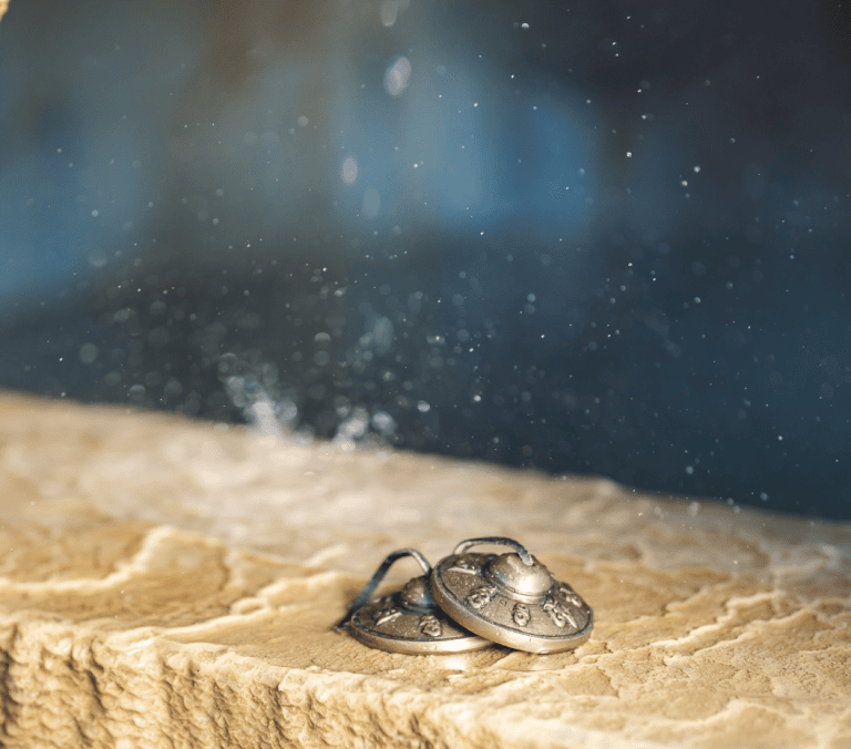Two ornate, bronze-colored circular objects, linked by a black cord, rest on a light brown, rough stone surface. Water drips from above, creating a fine mist against a blurred dark blue background.