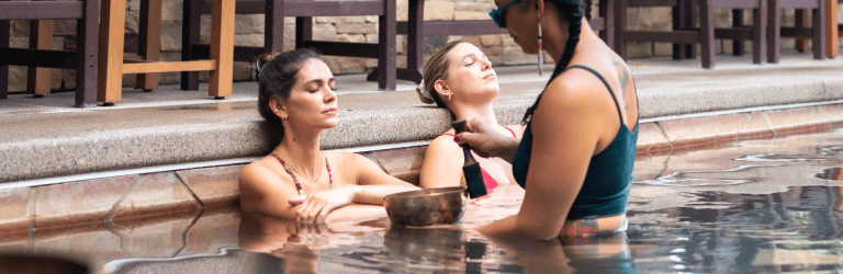 Three women in a pool. Two relax with closed eyes, one holding a singing bowl and mallet near another's head. Surrounded by tiled pool walls and wooden outdoor furniture.