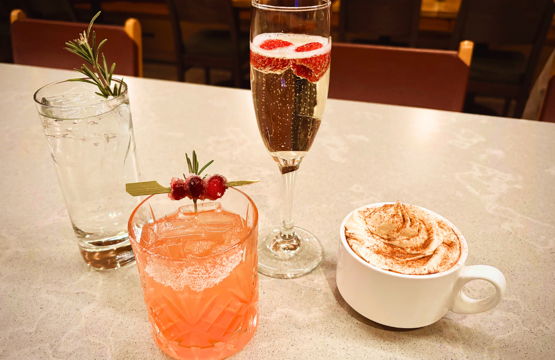 Four garnished beverages—water, a pink cocktail, sparkling wine, and coffee—are displayed on a speckled counter, with blurred restaurant seating in the background.