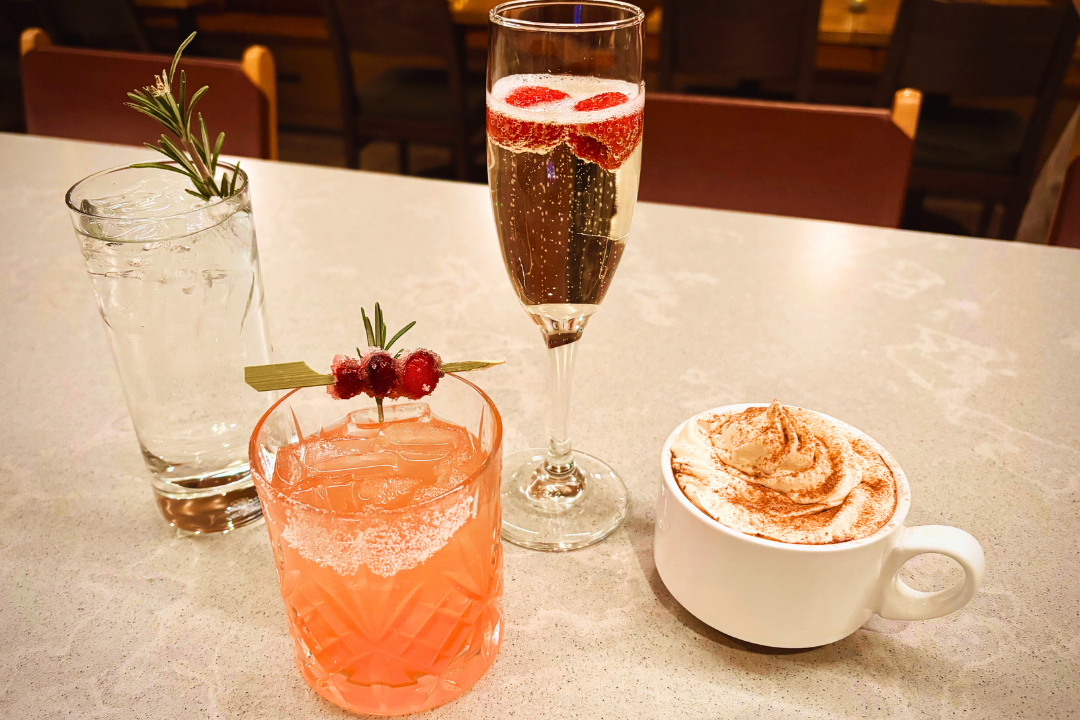 Four garnished beverages—water, a pink cocktail, sparkling wine, and coffee—are displayed on a speckled counter, with blurred restaurant seating in the background.