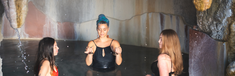 Three women are immersed in a cave hot spring. The center woman holds small cymbals, while the others relax with closed eyes. Water drips from rock formations.