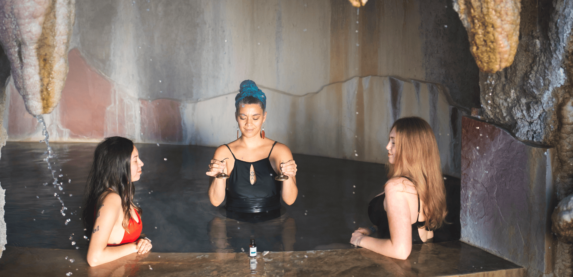 Three women are immersed in a cave hot spring. The center woman holds small cymbals, while the others relax with closed eyes. Water drips from rock formations.