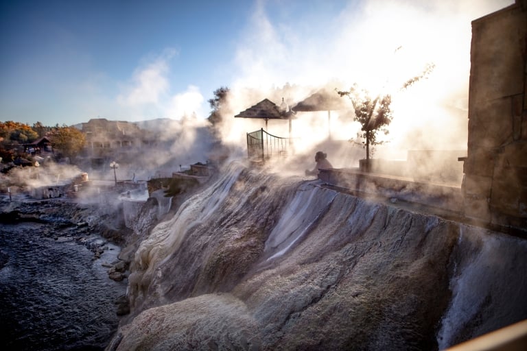 A person soaks in a steaming outdoor hot spring pool beside a river, with mineral-rich rock formations. Buildings and umbrellas are visible under a bright, hazy sky.