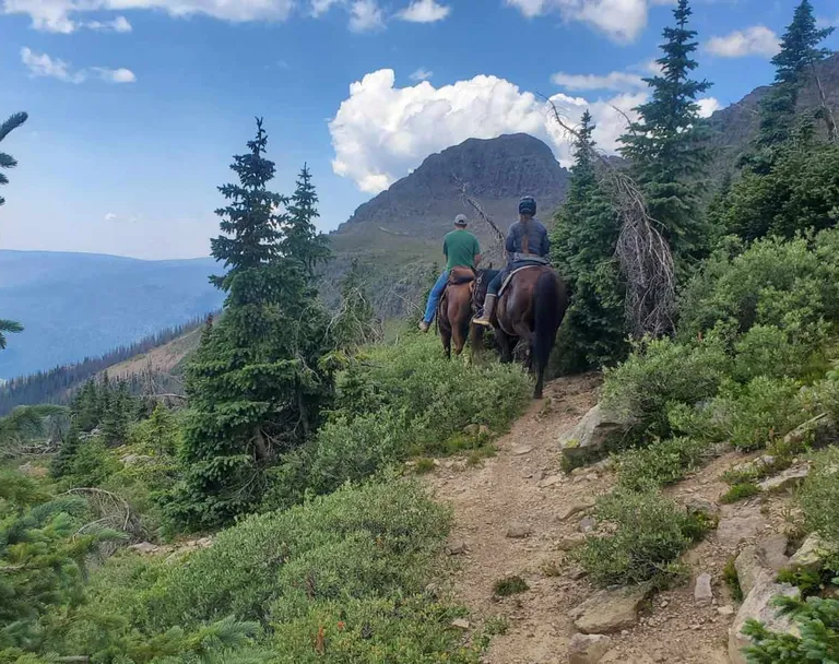 Two riders on horseback ascend a dirt trail through a coniferous mountain forest. A large peak rises under a cloudy sky in the background.