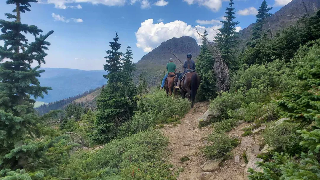 Two riders on horseback ascend a dirt trail through a coniferous mountain forest. A large peak rises under a cloudy sky in the background.