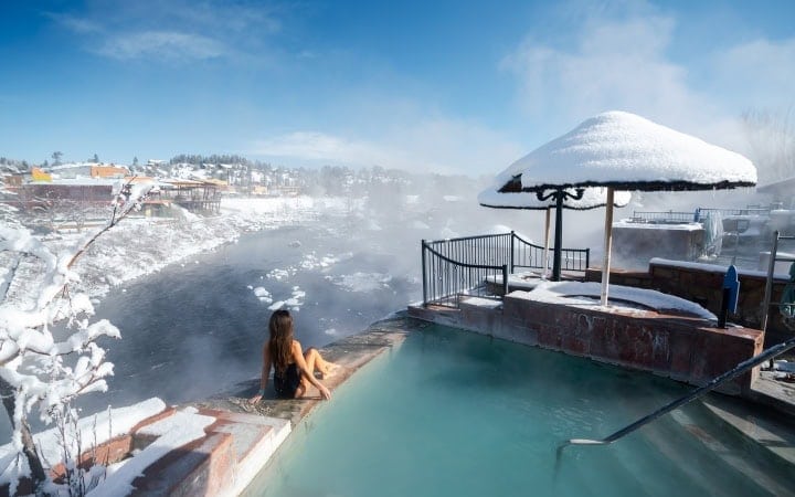 A woman sits by a steaming hot spring pool, overlooking a snowy river and town under a clear blue winter sky.