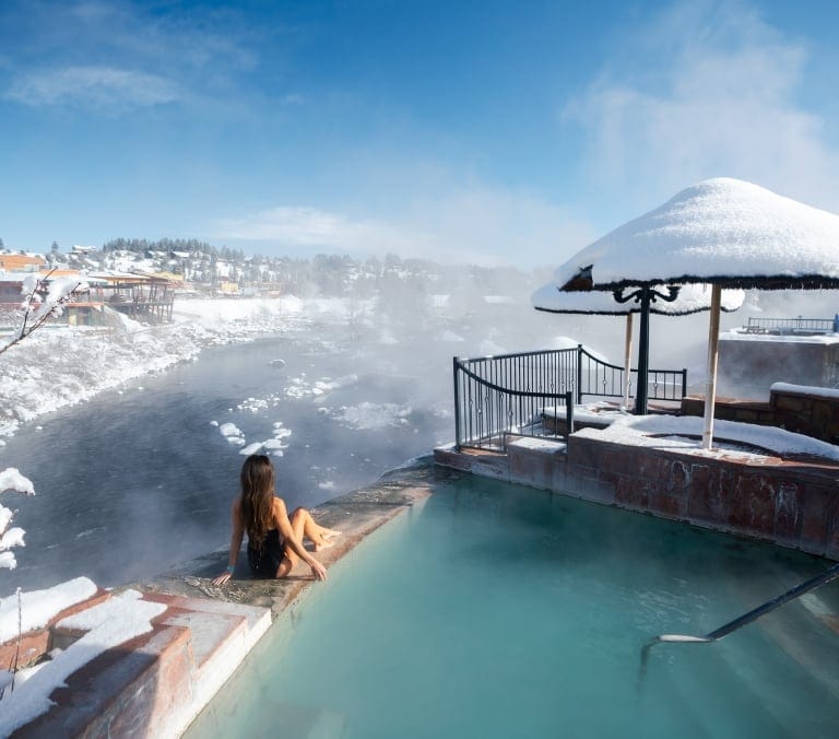 A woman sits by a steaming hot spring pool, overlooking a snowy river and town under a clear blue winter sky.