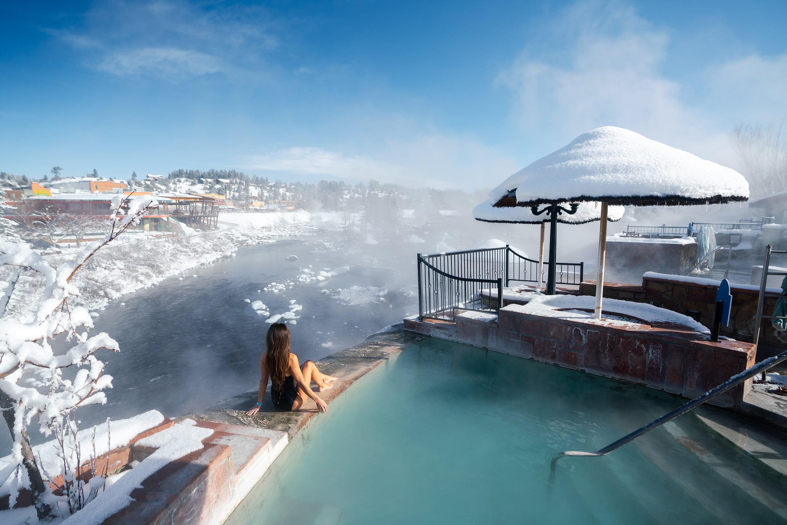 A woman sits by a steaming hot spring pool, overlooking a snowy river and town under a clear blue winter sky.