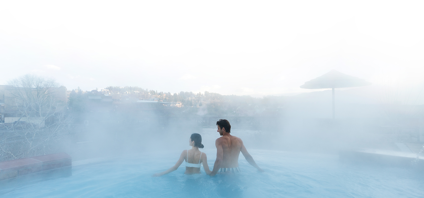 A man and woman stand waist-deep in a steaming blue outdoor pool, looking towards a distant town on a misty hillside.