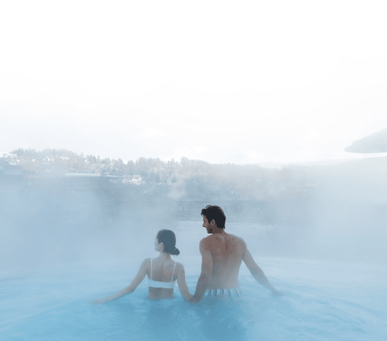 A couple stands with backs to camera in a steaming blue outdoor pool. They gaze at a hazy winter landscape with distant buildings and trees. A thatched umbrella is on the right.