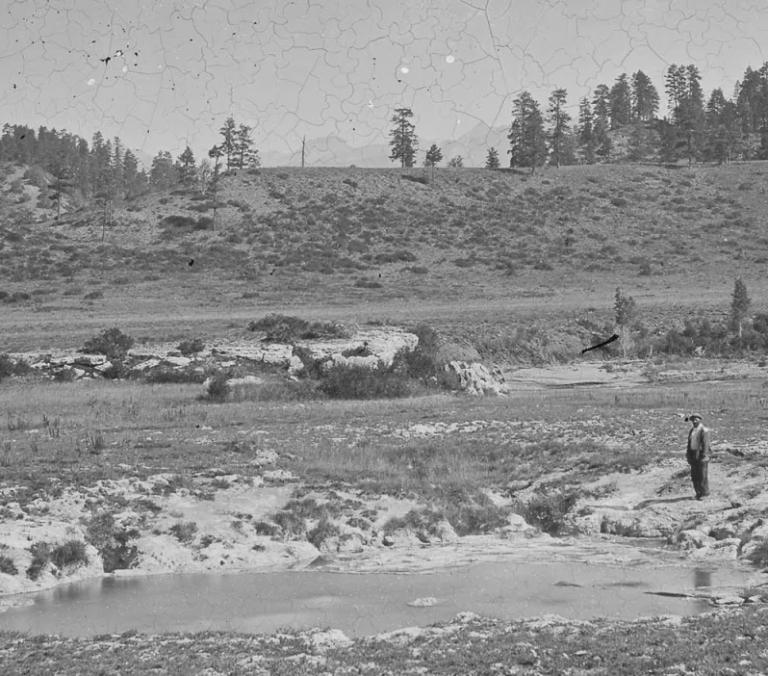 A man stands by a small pool in a desolate, rocky, grassy landscape with rolling, tree-covered hills under a hazy sky.