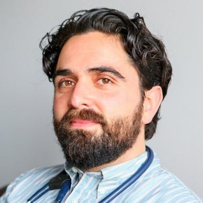 A bearded man with dark, curly hair gazes forward, slightly smiling, wearing a striped shirt and a blue stethoscope around his neck, against a plain gray background.