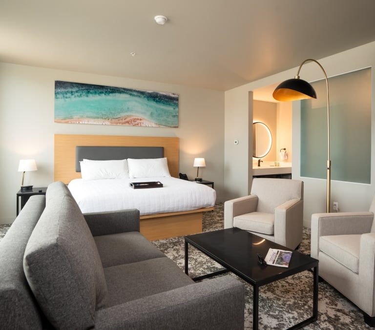 Modern hotel room: A king bed sits under an abstract painting, facing a gray sofa, two armchairs, and a coffee table. A magazine titled 'VIEW' rests on the table. A bathroom is visible.