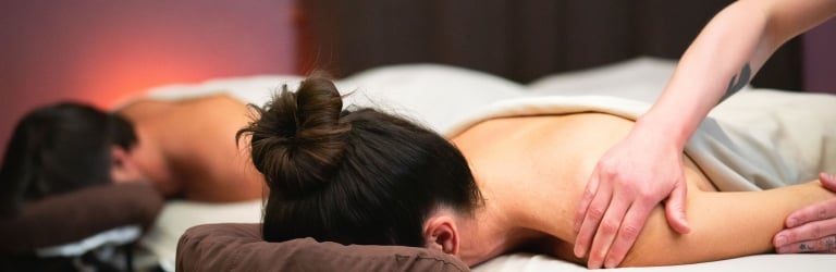 A woman lies face down on a massage table, receiving a shoulder and arm massage from tattooed hands. Another person is visible in the background in a dimly lit, warm-toned spa room.
