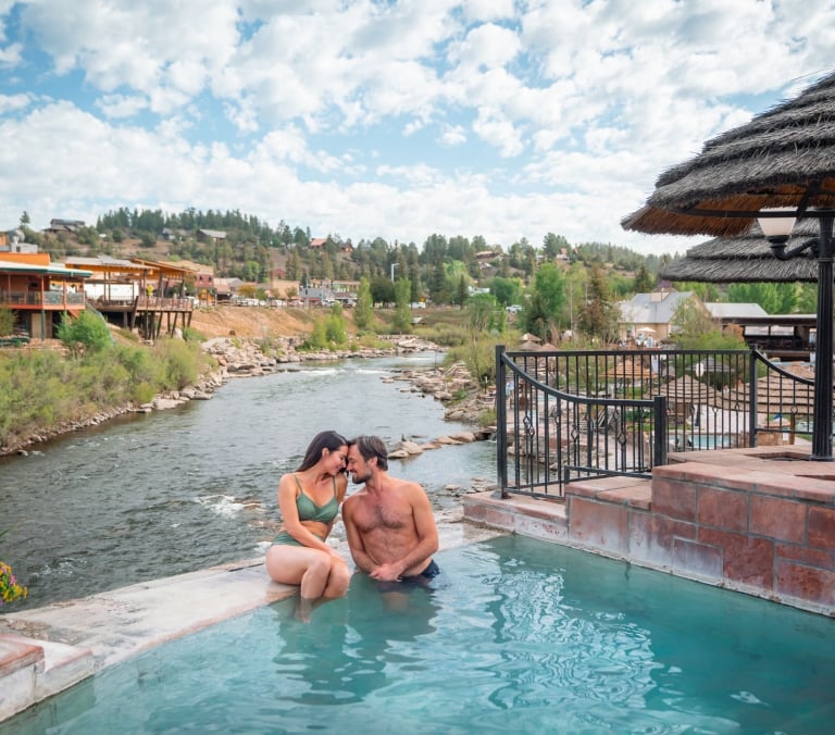 A couple embraces in a turquoise hot tub by a river. Beyond them, a town with buildings and forested hills sits under a cloudy sky.