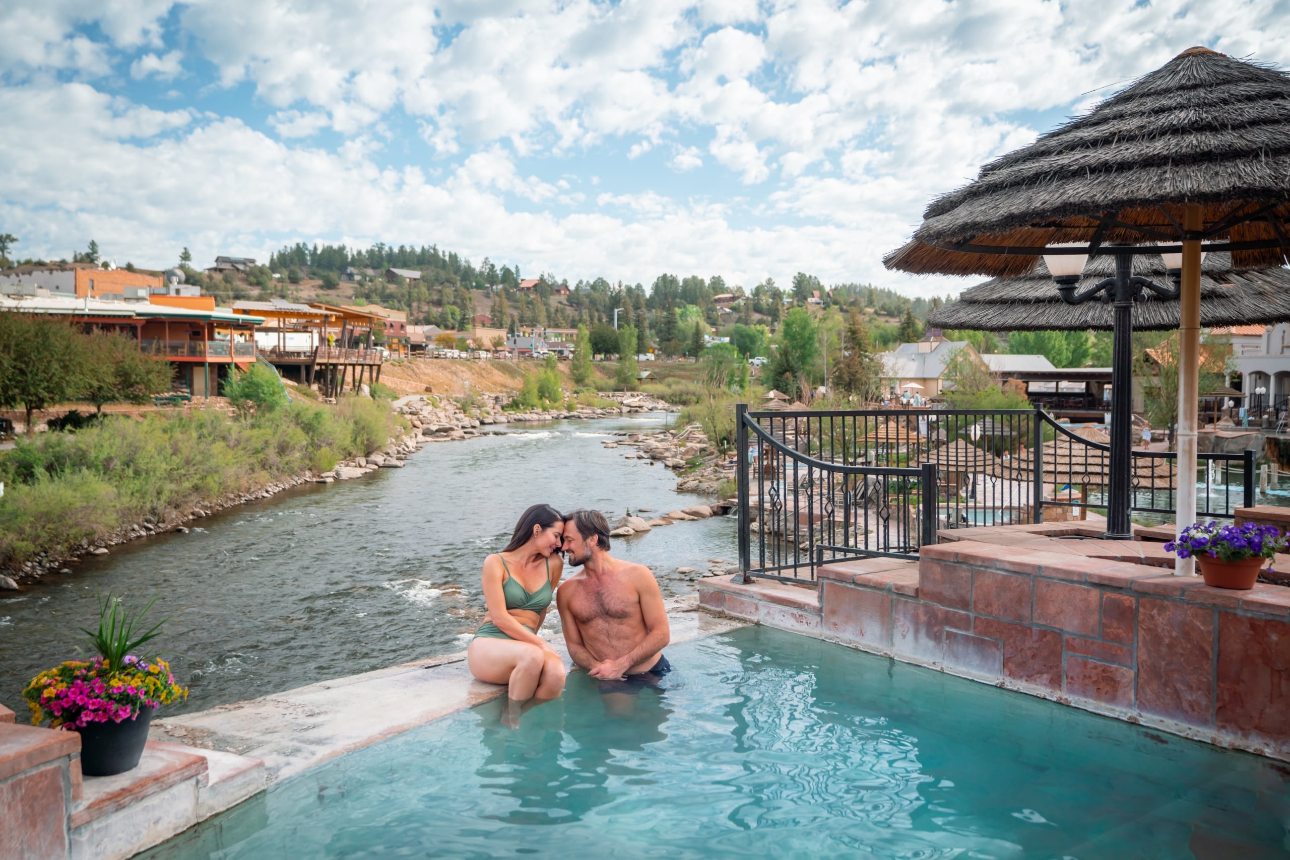 A couple embraces in a turquoise hot tub by a river. Beyond them, a town with buildings and forested hills sits under a cloudy sky.