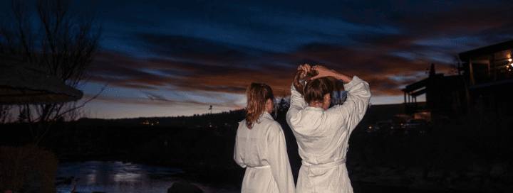 Two women in light robes stand by a river at dusk; one adjusts her hair. A colorful sky stretches above dark hills and distant buildings.