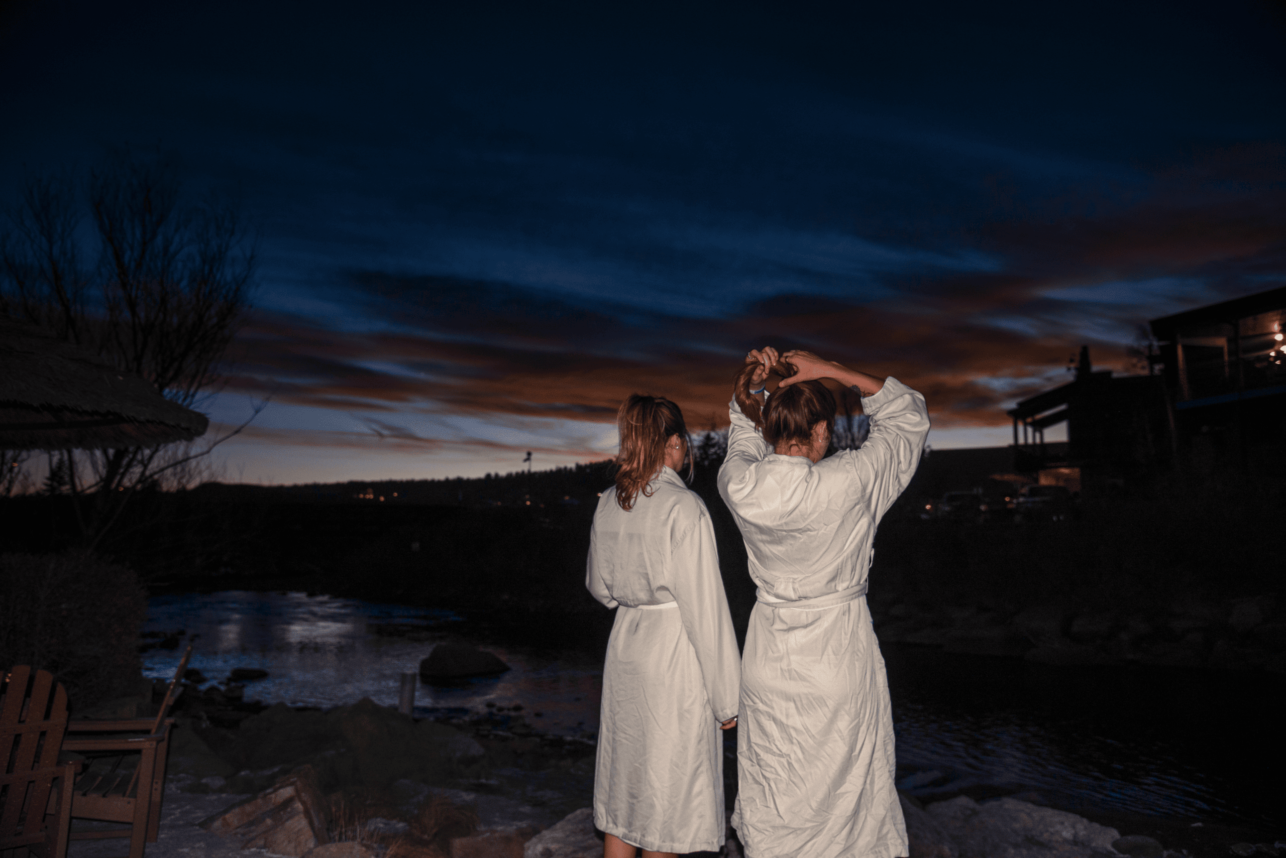 Two women in light robes stand by a river at dusk; one adjusts her hair. A colorful sky stretches above dark hills and distant buildings.