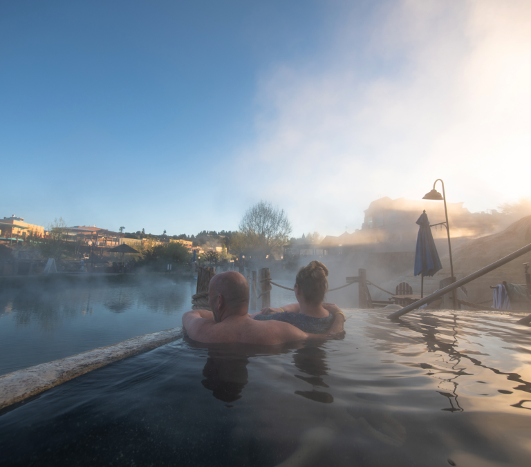 A couple relaxes in a steaming hot spring pool, facing a misty lake with resort buildings visible under a bright, clear sky.