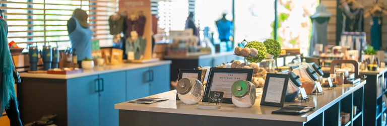 A modern retail store interior displays various lifestyle products including clothing, bath items, and packaged goods on multiple counters. Text: 