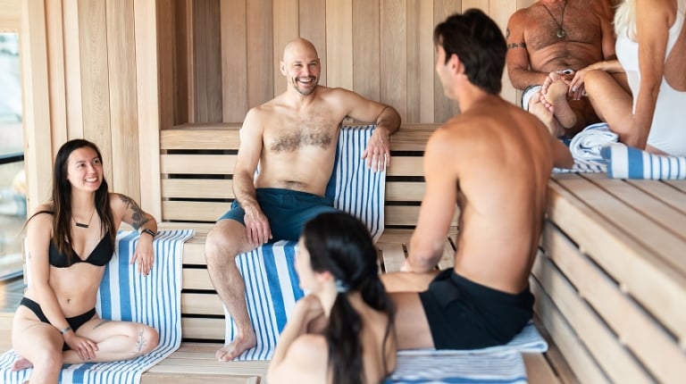 Six people in swim attire are sitting and conversing happily on wooden benches in a sauna, with striped towels beneath them.