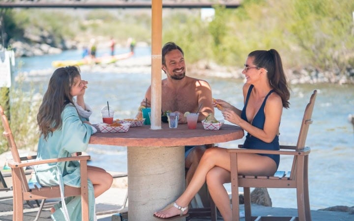 Two women and a man are eating snacks and conversing at a round table under an umbrella. They are next to a river with a bridge and distant watercraft.