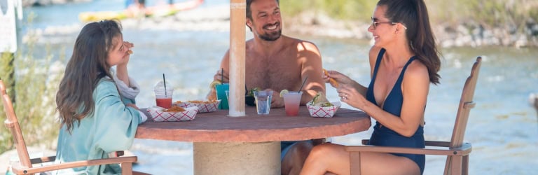 Two women and a man are eating snacks and conversing at a round table under an umbrella. They are next to a river with a bridge and distant watercraft.