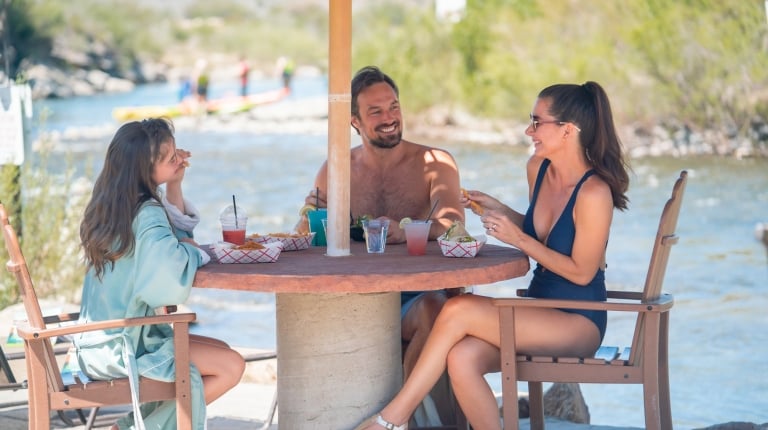 Two women and a man are eating snacks and conversing at a round table under an umbrella. They are next to a river with a bridge and distant watercraft.