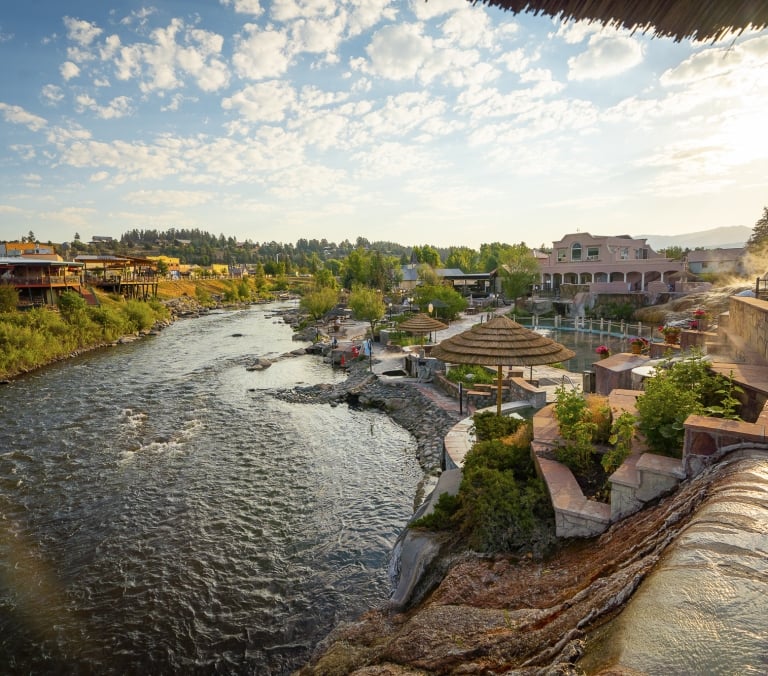 A vibrant river flows alongside a tiered hot springs resort with straw umbrellas and steaming pools. A colorful town and distant mountains frame the scene under a partly cloudy sky.