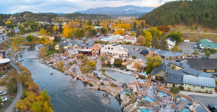 Aerial view: Hot springs complex with numerous pools where people relax, bordering a river and a vibrant town nestled among autumn trees and forested mountains.