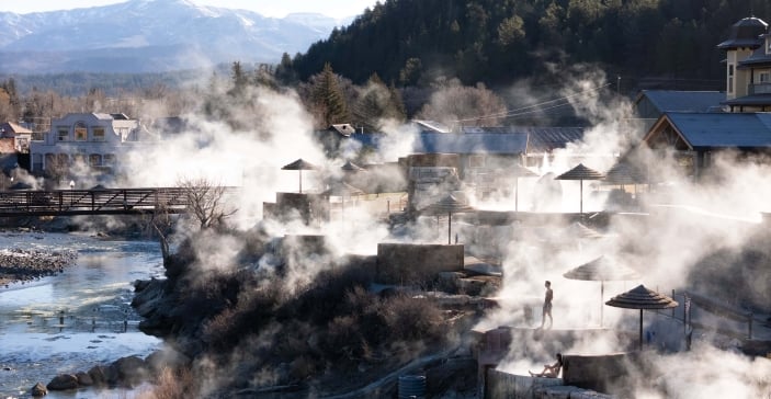 Steaming terraced hot springs with bathers (man standing, woman sitting) next to a river, bridge, buildings (one marked 'THE SPRINGS'), and distant snowy mountains.